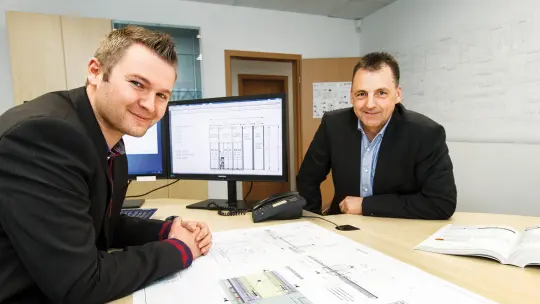 Two men sitting at the desk smiling in front of plans